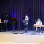 Muzikinis festivalis-konkursas "Muzikoje - mes šeima" ant scenos moteris groja pianinu, mergaitė groja smuiku, kita mergaitė groja kanklėmis