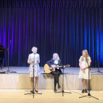 Muzikinis festivalis-konkursas "Muzikoje - mes šeima" trys žmonės atlieka kūrinį prie scenos, viena moteris su gitara