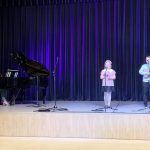 Muzikinis festivalis-konkursas "Muzikoje - mes šeima" ant scenos moteris groja pianinu, mergaitė dainuoja, berniukas groja gitarėle